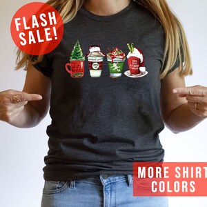 May include: Dark gray t-shirt featuring a festive graphic of four holiday-themed coffee drinks. The design includes a Christmas tree, Santa hat, and whipped cream. The shirt has the text "Flash Sale!" and "More Shirt Colors".