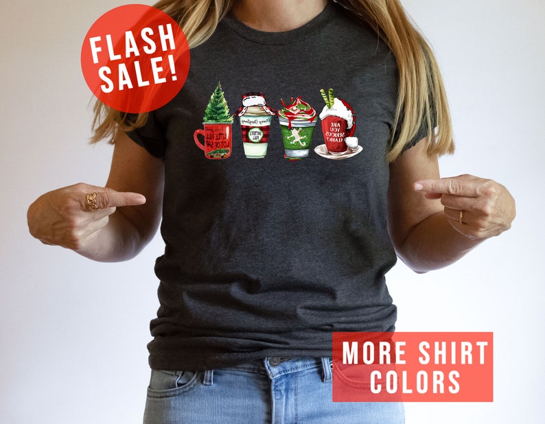 May include: Dark gray t-shirt featuring a festive graphic of four holiday-themed coffee drinks. The design includes a Christmas tree, Santa hat, and whipped cream. The shirt has the text "Flash Sale!" and "More Shirt Colors".