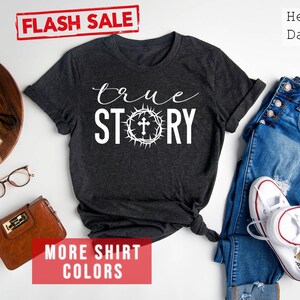 May include: A heather dark grey t-shirt with the text "true STORY" in white lettering. A crown of thorns with a cross is centered between the words "true" and "STORY".