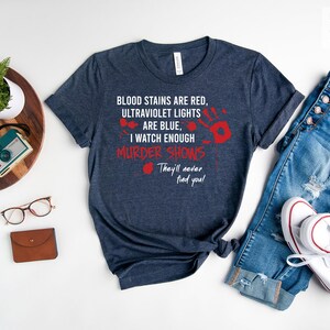 May include: A heather navy blue t-shirt with the text "BLOOD STAINS ARE RED, ULTRAVIOLET LIGHTS ARE BLUE, I WATCH ENOUGH MURDER SHOWS They'll never find you!" in white lettering with red blood splatter accents.
