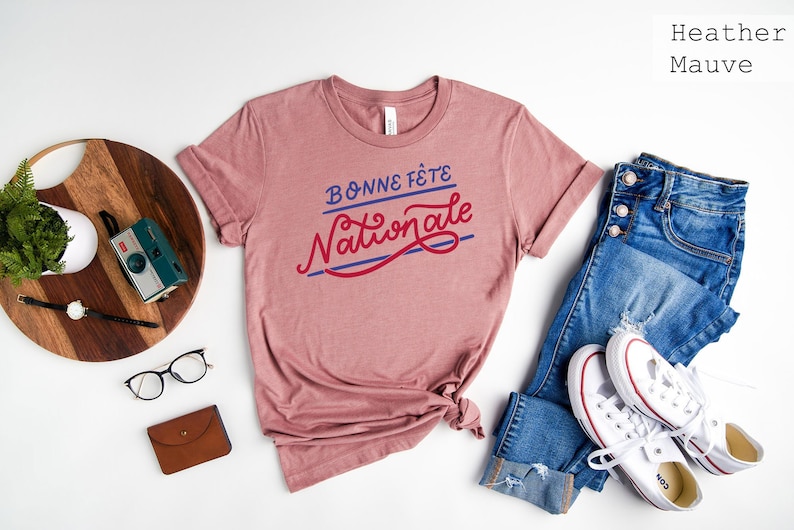 May include: A heather mauve short-sleeve t-shirt with the text "BONNE F&Ecirc;TE NATIONALE" printed in blue and red script. The shirt is paired with blue denim jeans and white canvas sneakers, accompanied by a camera, glasses, and a brown wallet.