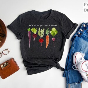 May include: Dark gray t-shirt with a watercolor illustration of various root vegetables. The shirt features the text "let's root for each other" in white. The shirt is paired with blue jeans and white sneakers.