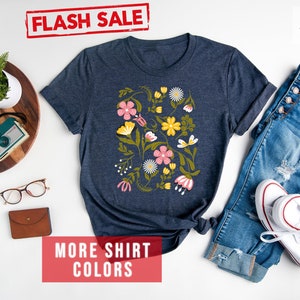 May include: A navy blue t-shirt with a floral print featuring pink, yellow, and green flowers. The shirt is displayed on a white background with a pair of jeans, white sneakers, and a wooden tray with a watch and a camera.
