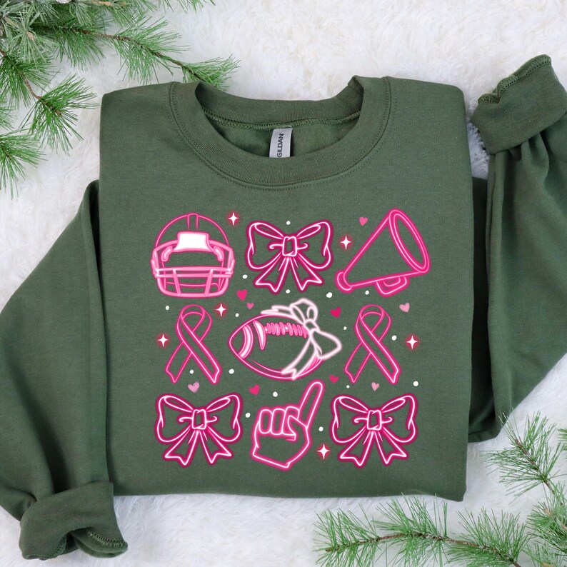 May include: A green sweatshirt with a pink design featuring a football, a megaphone, a ribbon, and bows. The design is reminiscent of a breast cancer awareness theme.