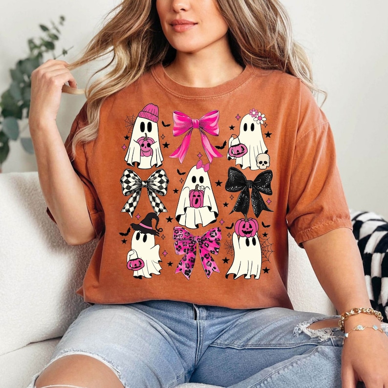 Puede incluir: Camiseta naranja quemado con fantasmas de dibujos animados con lazos y accesorios. Los fantasmas sostienen calabazas, usan sombreros y llevan bolsos. Los lazos son rosas, negros y a cuadros. Tambi&eacute;n se presentan estrellas negras y telara&ntilde;as.