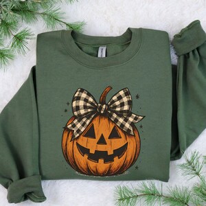 May include: A dark green sweatshirt featuring a smiling jack-o'-lantern with a black and white plaid bow. The pumpkin is orange with black details. The sweatshirt has long sleeves and a crew neck.