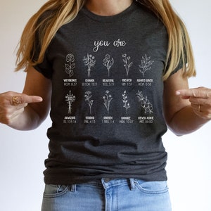 May include: A dark gray t-shirt with white floral designs and bible verses. The text reads "You are" and includes verses such as "Victorious, Rom 8:37", "Enough, 2 Cor 12:9", "Beautiful, Eccl 3:11", "Created, Gen 1:31", "Always Loved, Rom 8:38", "Amazing, Ps 139:14", "Strong, Phil 4:13", "Chosen, 1 Thess 1:4", "Capable, Mark 10:27", and "Never Alone, Matt 28:20".