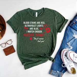 May include: A forest green t-shirt with white text that reads "Blood stains are red, ultraviolet lights are blue, I watch enough murder shows. They'll never find you!" The text is surrounded by red handprints.