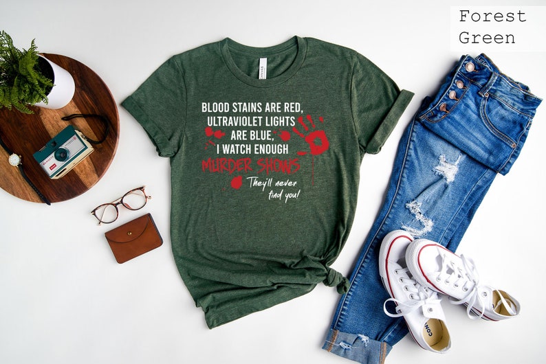 May include: A forest green t-shirt with white text that reads "Blood stains are red, ultraviolet lights are blue, I watch enough murder shows. They'll never find you!" The text is surrounded by red handprints.
