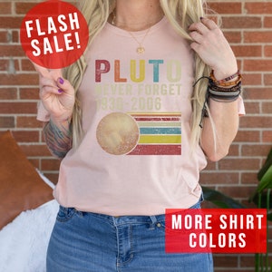 Op de afbeelding: Een roze T-shirt met een retro-stijl grafische afbeelding met de tekst "Pluto Never Forget 1930-2006" en een gestileerde afbeelding van Pluto. De afbeelding heeft een rood, geel, blauw en groen kleurenschema.