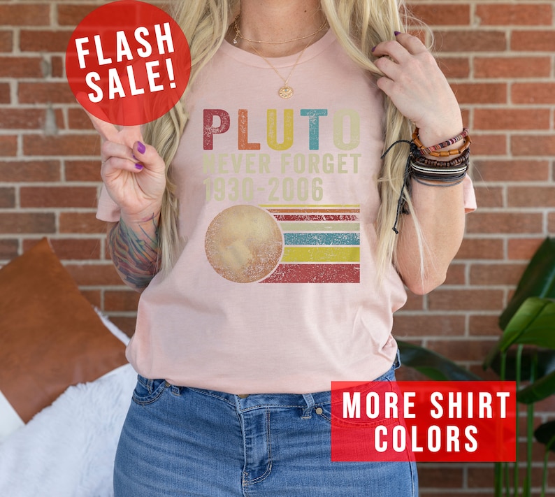 Op de afbeelding: Een roze T-shirt met een retro-stijl grafische afbeelding met de tekst "Pluto Never Forget 1930-2006" en een gestileerde afbeelding van Pluto. De afbeelding heeft een rood, geel, blauw en groen kleurenschema.