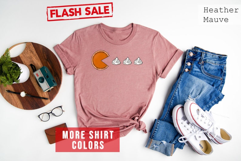 K&ouml;nnte beinhalten: Ein T-Shirt in Heather Mauve mit einem Aufdruck einer K&uuml;rbis-Tortenst&uuml;ck, das drei Kleckse Schlagsahne isst. Der Text "FLASH SALE" ist in Rot &uuml;ber dem Shirt. Der Text "MORE SHIRT COLORS" ist in Rot unter dem Shirt.