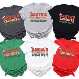 Puede incluir: Seis camisetas de manga corta en varios colores: carbón, gris, negro, verde, blanco y rojo. Cada camiseta presenta el texto "SANTA'S FAVORITE OFFICE STAFF" en una fuente retro con un gorro de Papá Noel, ideal para la oficina en Navidad.