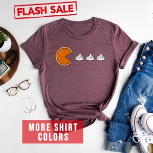 K&ouml;nnte beinhalten: Ein T-Shirt in Heather-Maroon mit einem Grafikdruck von Pac-Man, der drei Mini-K&uuml;rbiskuchen isst. Der Text "FLASH SALE" ist oben im Bild in Rot. Der Text "MORE SHIRT COLORS" ist unten im Bild in Rot.