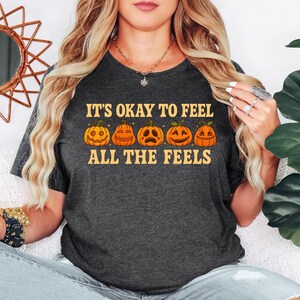 May include: Dark gray t-shirt with the text "IT'S OKAY TO FEEL ALL THE FEELS" in orange. Below the text are five orange jack-o'-lanterns with different facial expressions. The shirt is a crew neck and is made of a soft material.