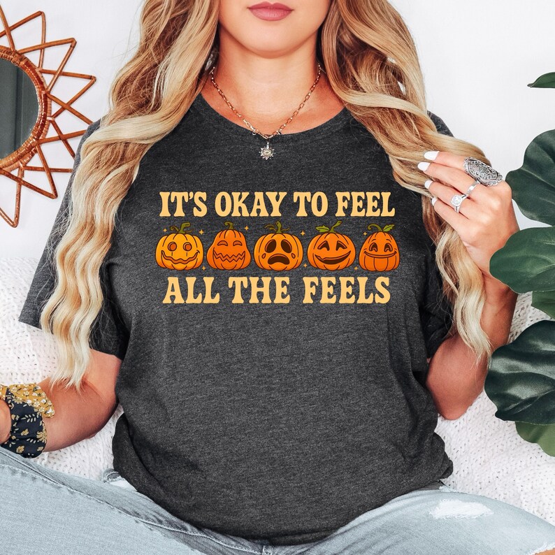 May include: Dark gray t-shirt with the text "IT'S OKAY TO FEEL ALL THE FEELS" in orange. Below the text are five orange jack-o'-lanterns with different facial expressions. The shirt is a crew neck and is made of a soft material.