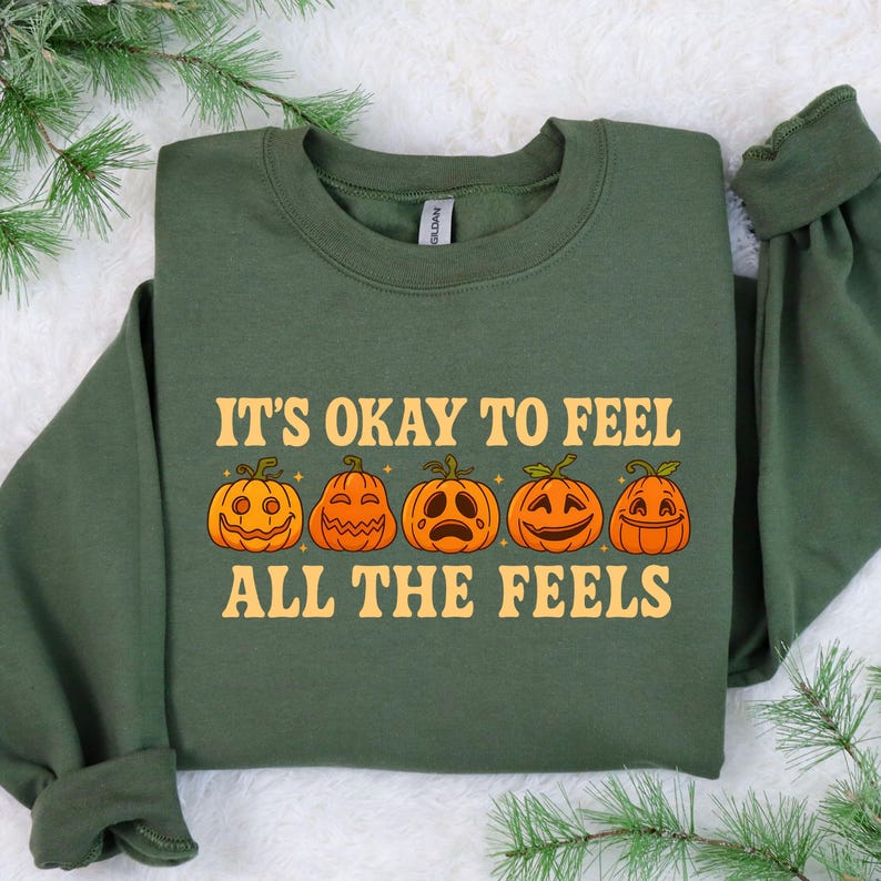 May include: Green sweatshirt with the text "IT'S OKAY TO FEEL ALL THE FEELS" and five orange jack-o'-lanterns with different facial expressions. The sweatshirt has long sleeves and a crew neck. Perfect for Halloween or fall.
