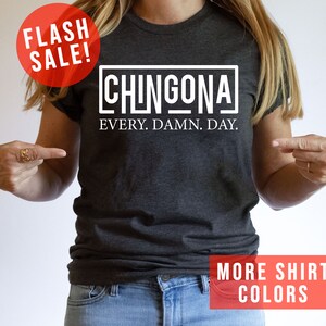May include: A dark gray t-shirt with white text that reads "CHINGONA EVERY. DAMN. DAY." The model is wearing the shirt and pointing to it.