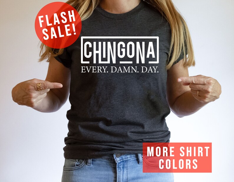 May include: A dark gray t-shirt with white text that reads "CHINGONA EVERY. DAMN. DAY." The model is wearing the shirt and pointing to it.