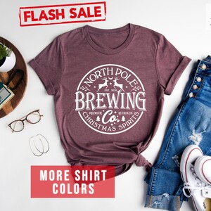 May include: Heather maroon t-shirt with a white circular graphic that reads "North Pole Brewing Co. Premium Authentic Christmas Spirits." The shirt is displayed with blue jeans and white sneakers. The words "Flash Sale" and "More Shirt Colors" are also visible.