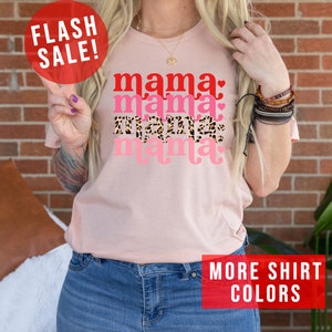 Op de afbeelding: Roze T-shirt met de tekst "mama" die vier keer wordt herhaald in verschillende kleuren en patronen. De tekst is in een retro-lettertype. Het shirt wordt gedragen door een persoon met een tatoeage op hun arm. De tekst "FLASH SALE!" staat in een rode cirkel linksboven in de afbeelding. De tekst "MORE SHIRT COLORS" staat in een rode rechthoek rechtsonder in de afbeelding.