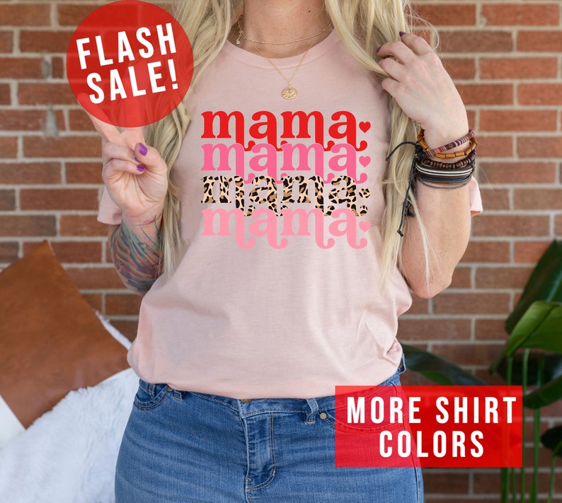 Op de afbeelding: Roze T-shirt met de tekst "mama" die vier keer wordt herhaald in verschillende kleuren en patronen. De tekst is in een retro-lettertype. Het shirt wordt gedragen door een persoon met een tatoeage op hun arm. De tekst "FLASH SALE!" staat in een rode cirkel linksboven in de afbeelding. De tekst "MORE SHIRT COLORS" staat in een rode rechthoek rechtsonder in de afbeelding.