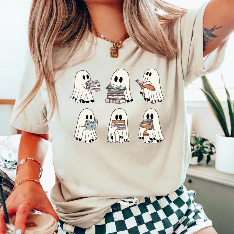 K&ouml;nnte beinhalten: Beiges T-Shirt mit einer Grafik von sechs Cartoon-Geistern, die jeweils B&uuml;cher halten. Die Geister sind wei&szlig; mit schwarzen Augen, und die B&uuml;cher sind in verschiedenen Farben. Das T-Shirt hat einen runden Ausschnitt und kurze &Auml;rmel.