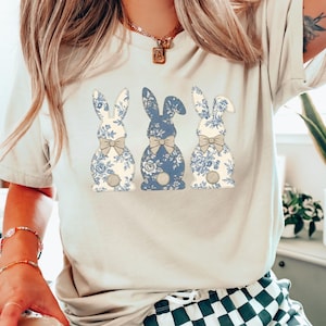 Puede incluir: Camiseta beige claro con tres diseños de conejos. Cada conejo tiene un estampado floral en azul y blanco, con una pajarita gris. El conejo del medio es azul liso. El diseño está centrado en la camiseta.