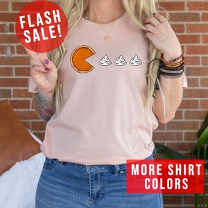 K&ouml;nnte beinhalten: Ein pinkfarbenes T-Shirt mit einem Aufdruck einer K&uuml;rbis-Tortenst&uuml;ckes, das Schlagsahne isst. Der Text "FLASH SALE!" befindet sich in einem roten Kreis &uuml;ber dem Aufdruck. Der Text "MORE SHIRT COLORS" befindet sich in einem roten Rechteck am unteren Rand des Bildes.
