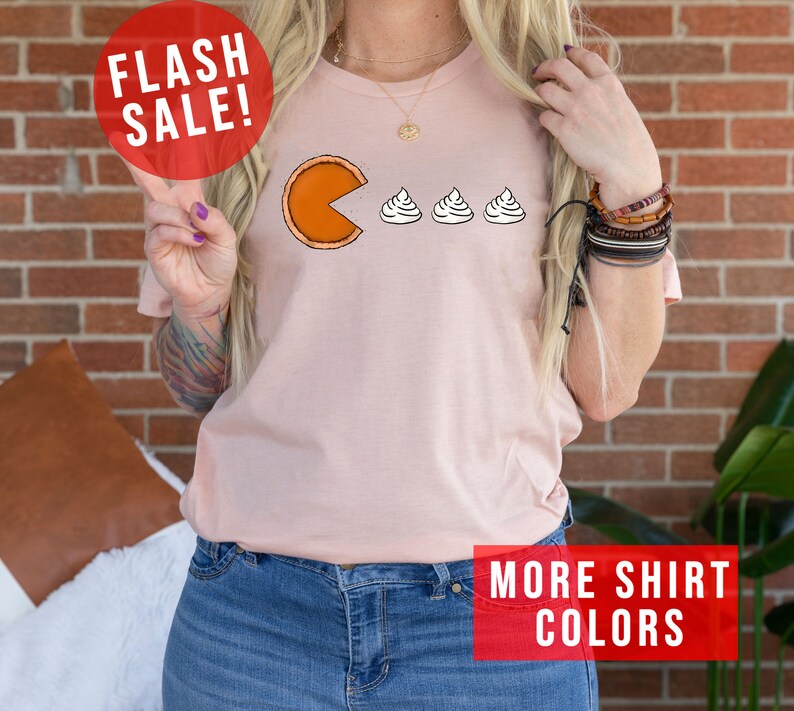 K&ouml;nnte beinhalten: Ein pinkfarbenes T-Shirt mit einem Aufdruck einer K&uuml;rbis-Tortenst&uuml;ckes, das Schlagsahne isst. Der Text "FLASH SALE!" befindet sich in einem roten Kreis &uuml;ber dem Aufdruck. Der Text "MORE SHIRT COLORS" befindet sich in einem roten Rechteck am unteren Rand des Bildes.