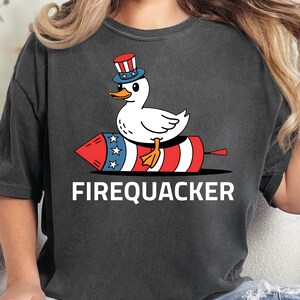 Peut inclure: T-shirt gris fonc&eacute; avec un canard de dessin anim&eacute; portant un chapeau patriotique, perch&eacute; sur un feu d'artifice rouge, blanc et bleu. Le texte "FIREQUACKER" est imprim&eacute; en dessous. Le t-shirt est de style d&eacute;contract&eacute;.