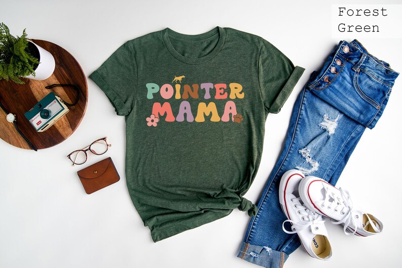May include: Forest green t-shirt featuring the text "POINTER MAMA" in colourful retro lettering. The shirt is styled with blue jeans and white trainers.