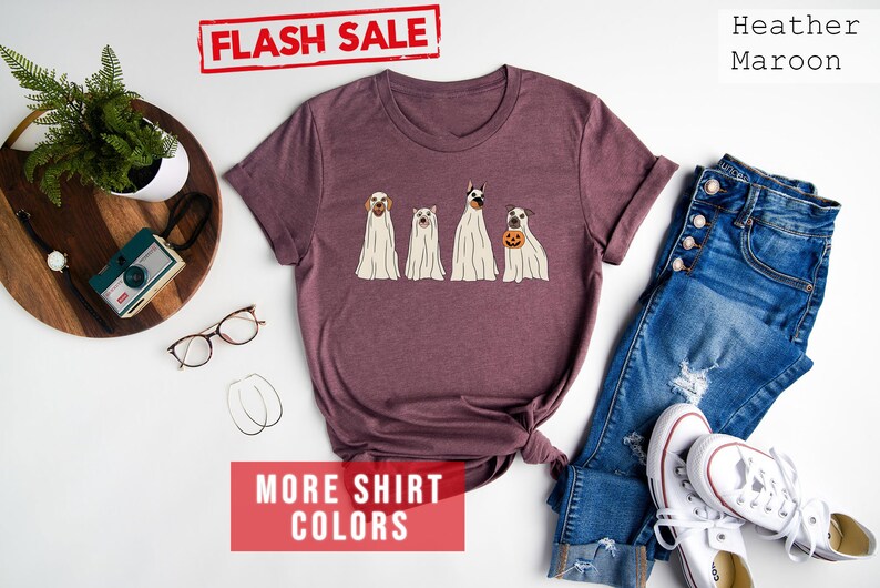 Op de afbeelding: Een heather maroon t-shirt met korte mouwen en een afbeelding van vier honden in witte spookkostuums, waarbij &eacute;&eacute;n hond een kleine oranje pompoen vasthoudt. Het shirt bevat de tekst FLASH SALE, Heather Maroon en MORE SHIRT COLORS.