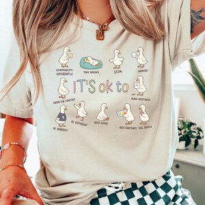 May include: A beige t-shirt with a graphic of cartoon ducks and the phrase "It's ok to." The ducks are shown in different situations, including taking breaks and avoiding eye contact. The shirt is a casual wear item.