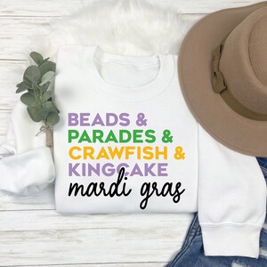 May include: White sweatshirt with the text "BEADS & PARADES & CRAWFISH & KINGCAKE mardi gras" in purple, green, yellow, and white lettering.