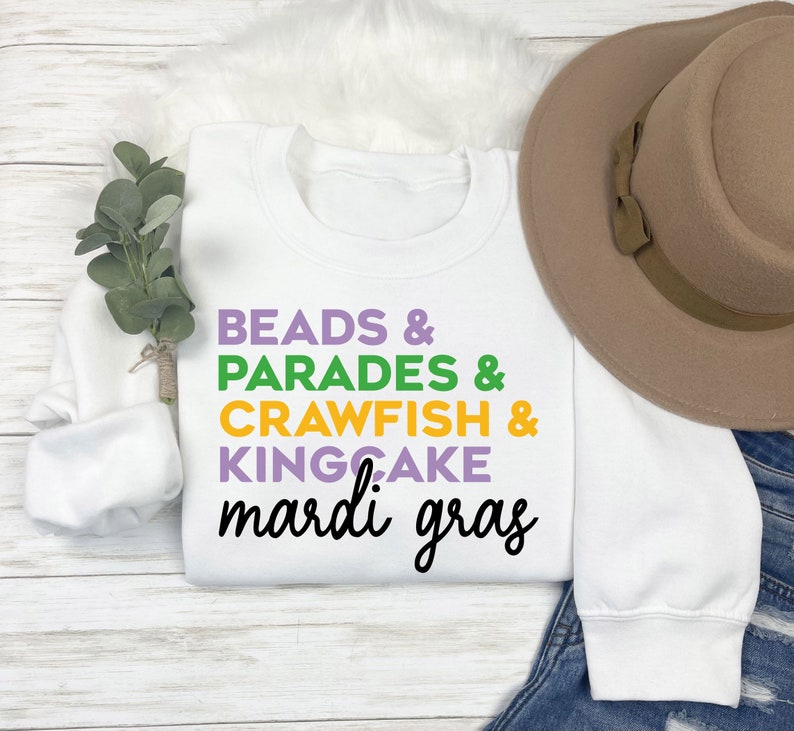 May include: White sweatshirt with the text "BEADS & PARADES & CRAWFISH & KINGCAKE mardi gras" in purple, green, yellow, and white lettering.