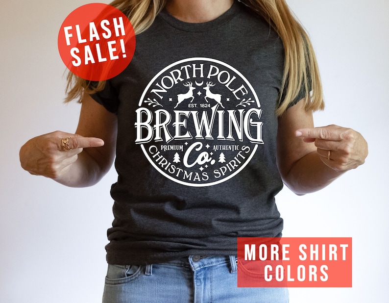 May include: Dark gray t-shirt with a white circular graphic that reads "North Pole Brewing Co. Premium Authentic Christmas Spirits." The shirt also has the text "Flash Sale!" and "More Shirt Colors" in red.