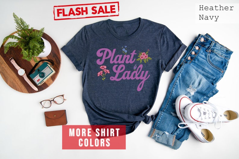 May include: A heather navy blue t-shirt with the text "Plant Lady" in purple script with floral accents. The shirt is laid flat on a white surface with a pair of jeans and white sneakers.