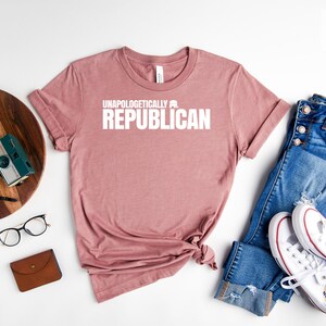 May include: Heather mauve t-shirt with the text "Unapologetically Republican" printed in white. The shirt is laid flat on a white surface with a pair of blue jeans, white sneakers, a wooden tray with a plant and a camera, and a brown leather wallet.