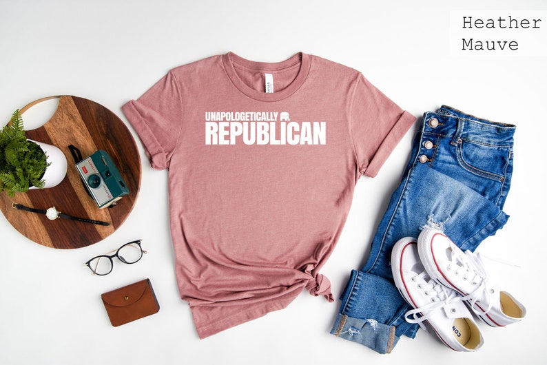 May include: Heather mauve t-shirt with the text "Unapologetically Republican" printed in white. The shirt is laid flat on a white surface with a pair of blue jeans, white sneakers, a wooden tray with a plant and a camera, and a brown leather wallet.