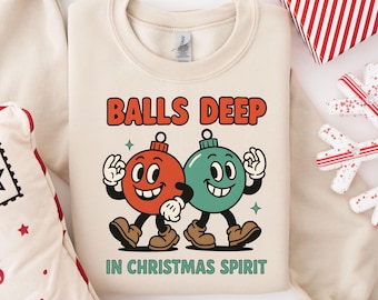 May include: Cream-colored sweatshirt with the text "BALLS DEEP IN CHRISTMAS SPIRIT." The graphic features two cartoon Christmas ornaments with faces, arms, and legs. One ornament is red, the other is teal.