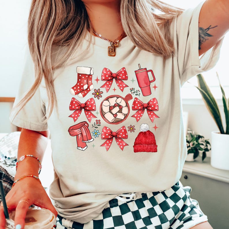 May include: Beige t-shirt featuring a festive graphic with red bows, a stocking, a scarf, a mug, and a beanie. The design includes a hot chocolate with marshmallows and floral accents, creating a cozy, holiday theme.