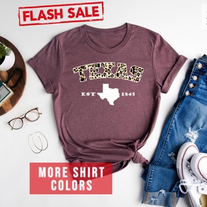 May include: A heather maroon t-shirt with a white graphic of the state of Texas with the word "TEXAS" in leopard print letters above it and "EST 1845" below it.