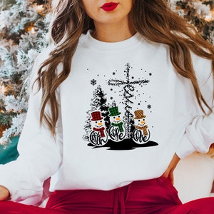 May include: White sweatshirt featuring a graphic of three snowmen with hats and scarves, and the words "Faith, Hope, Love". The design includes a cross and Christmas trees. The sweatshirt is a festive holiday item.