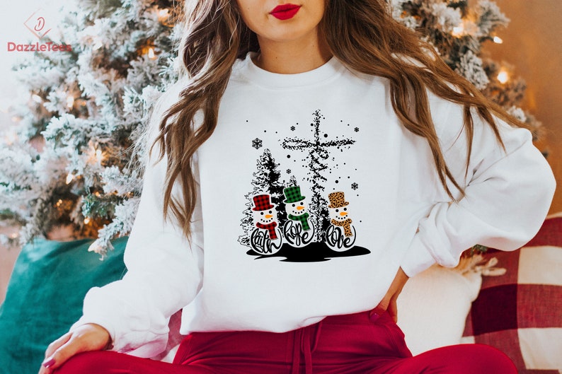 May include: White sweatshirt featuring a graphic of three snowmen with hats and scarves, and the words "Faith, Hope, Love". The design includes a cross and Christmas trees. The sweatshirt is a festive holiday item.