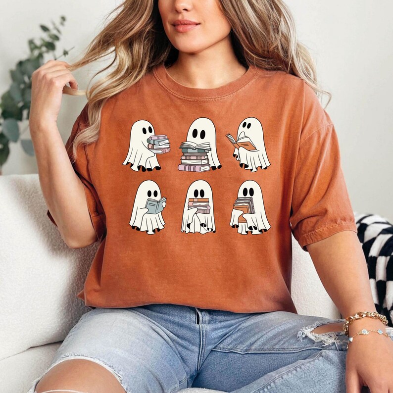 K&ouml;nnte beinhalten: Ein orangefarbenes T-Shirt mit einem Aufdruck von sechs Cartoon-Geistern, die jeweils B&uuml;cher halten oder lesen. Die Geister sind wei&szlig; mit schwarzen Augen und werden in verschiedenen Posen dargestellt. Das Shirt besteht aus einem weichen Material.
