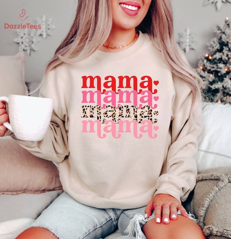 Op de afbeelding: Een cr&egrave;mekleurig sweatshirt met de tekst "mama" die vier keer wordt herhaald in rood, roze en luipaardprint. De tekst is in een retro-lettertype met harten.