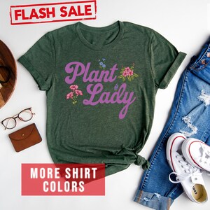 May include: A forest green t-shirt with the text "Plant Lady" in purple script with pink floral accents. The shirt is laid flat on a white surface with a pair of jeans and white sneakers.