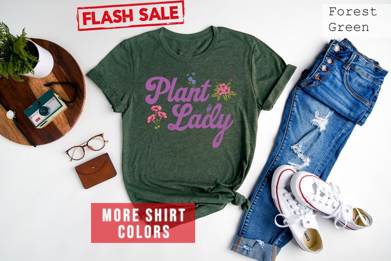 May include: A forest green t-shirt with the text "Plant Lady" in purple script with pink floral accents. The shirt is laid flat on a white surface with a pair of jeans and white sneakers.