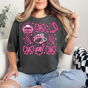 May include: A black t-shirt with a pink neon design featuring a football, a megaphone, a ribbon, and bows.